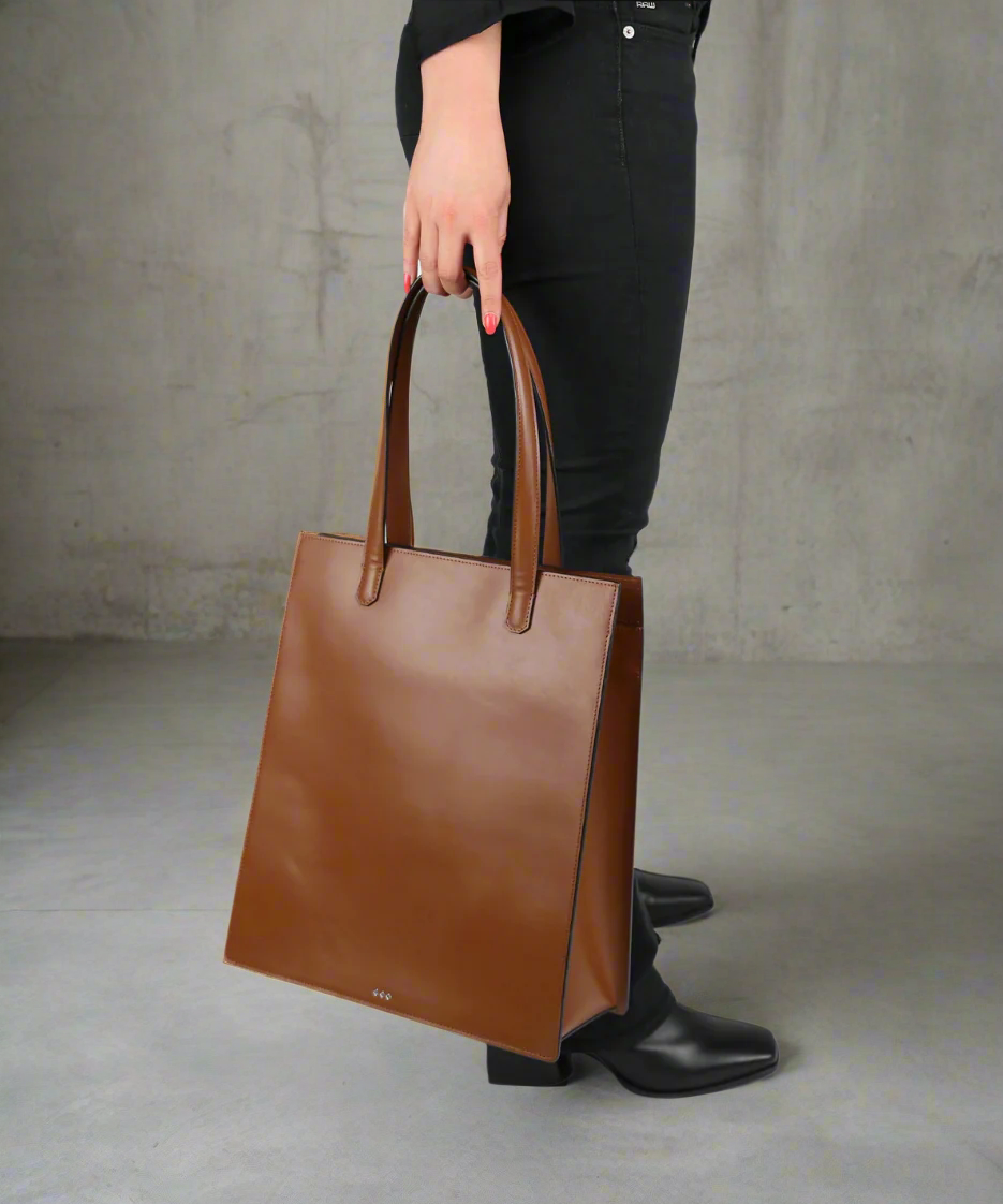 Brown leather tote bag held by a person wearing black pants and shoes on a grey background