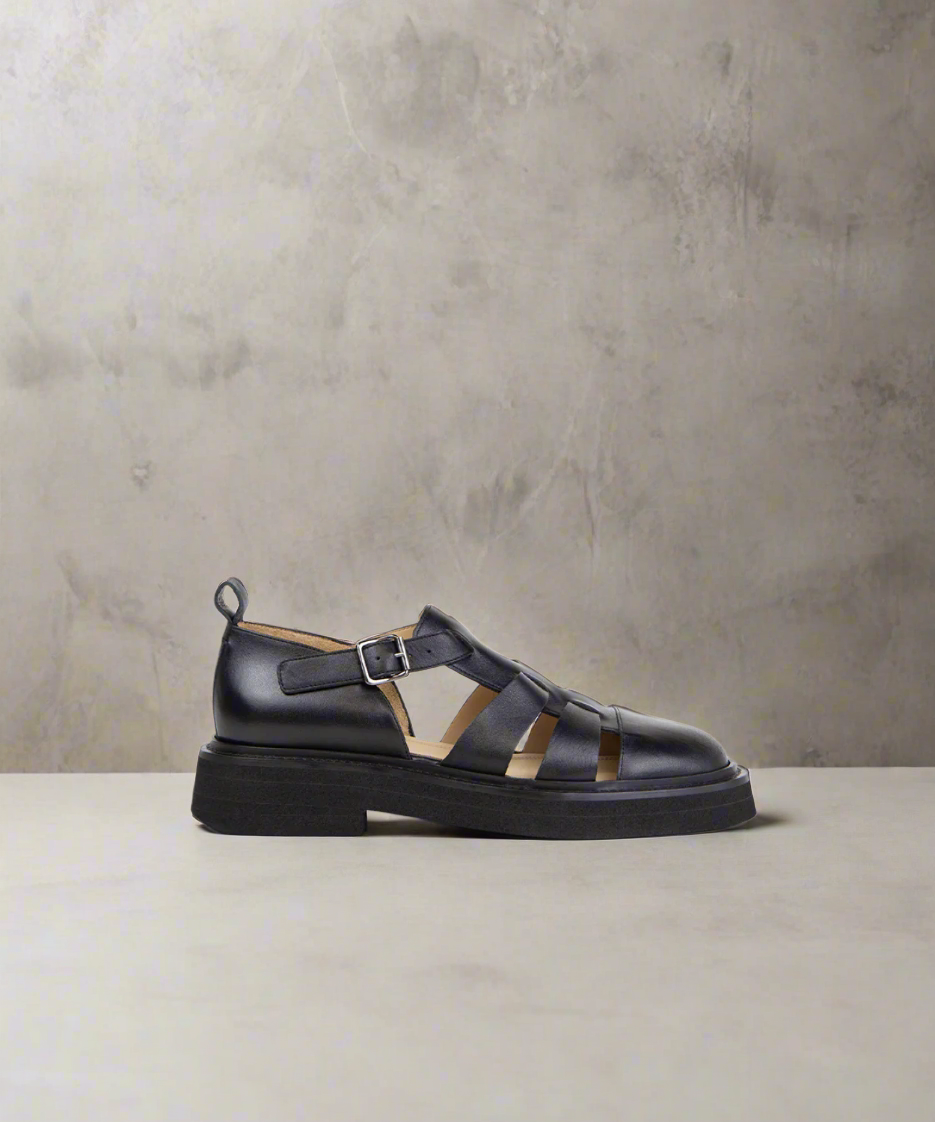 A black suede gladiator sandal for men with a metal buckle closure and cut-out detailing.