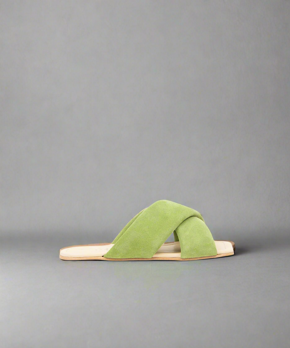 A green suede sandal with a flat heel and a curved toe line, displayed against a grey background.