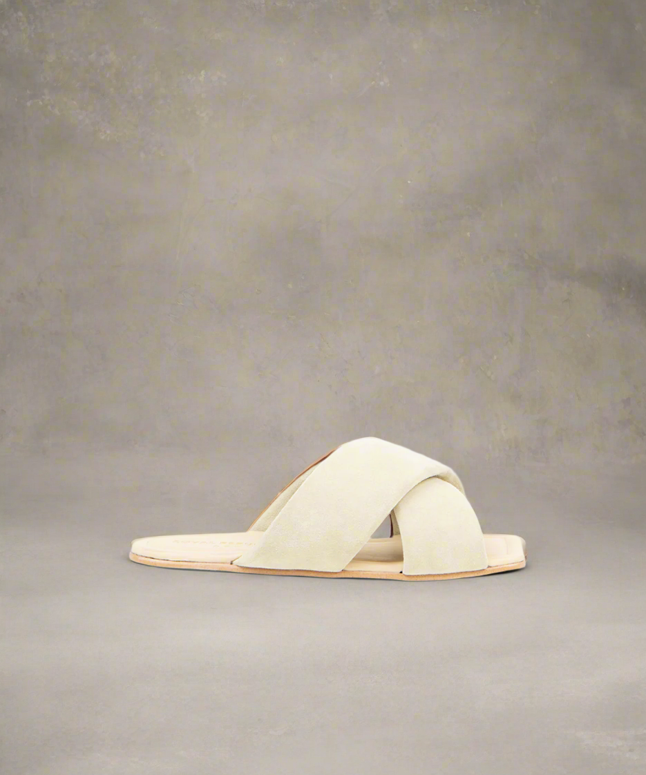 An off white suede sandal with a flat heel and a crisscross strap design, set against a grey background.