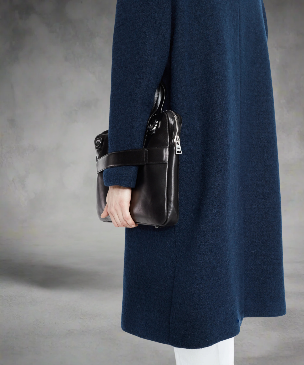 Person wearing a long navy coat holding a black Royal RepuliQ leather bag against a gray background
