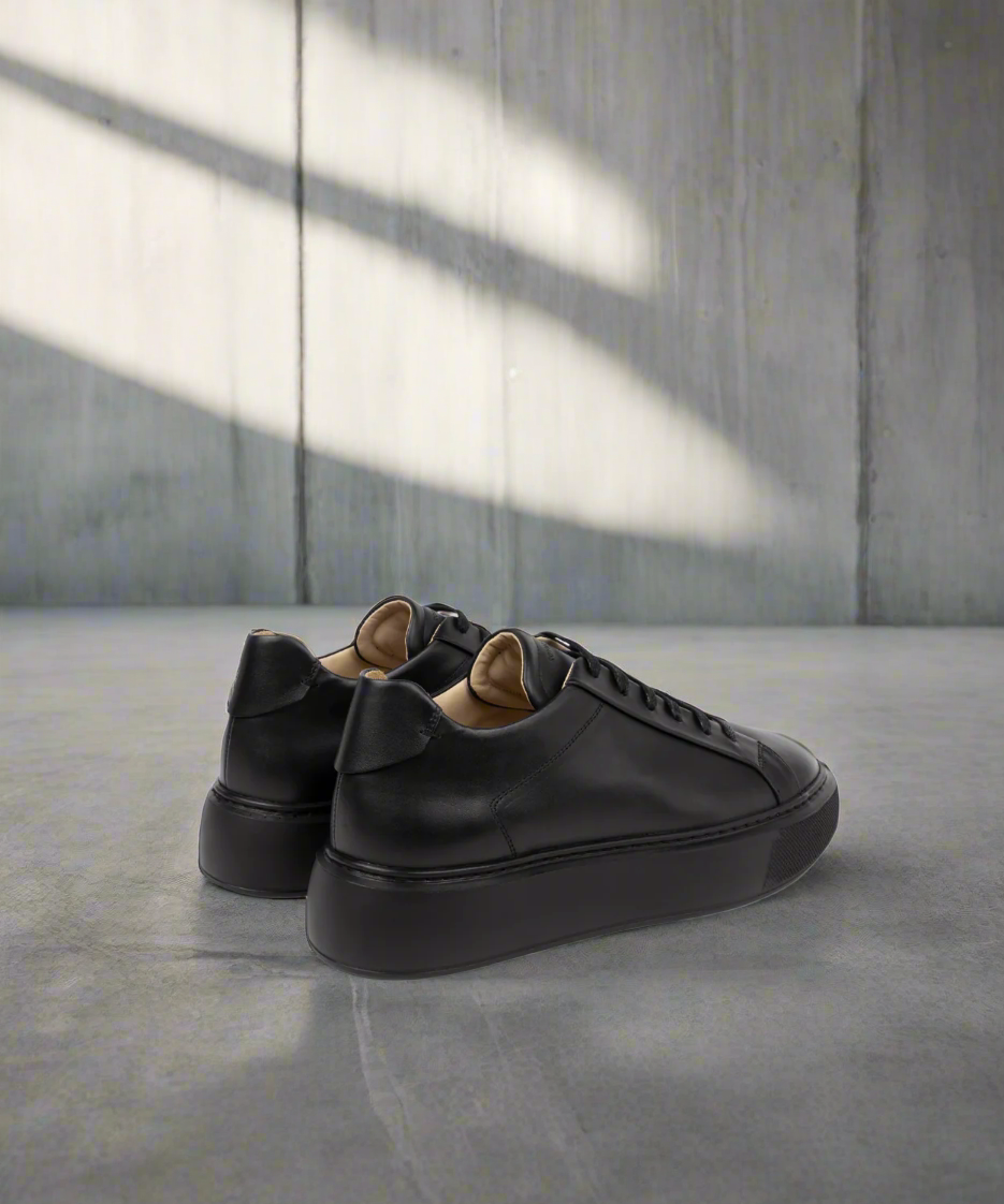 Pair of black Royal RepubliQ sneakers on a concrete floor with a gray wall background