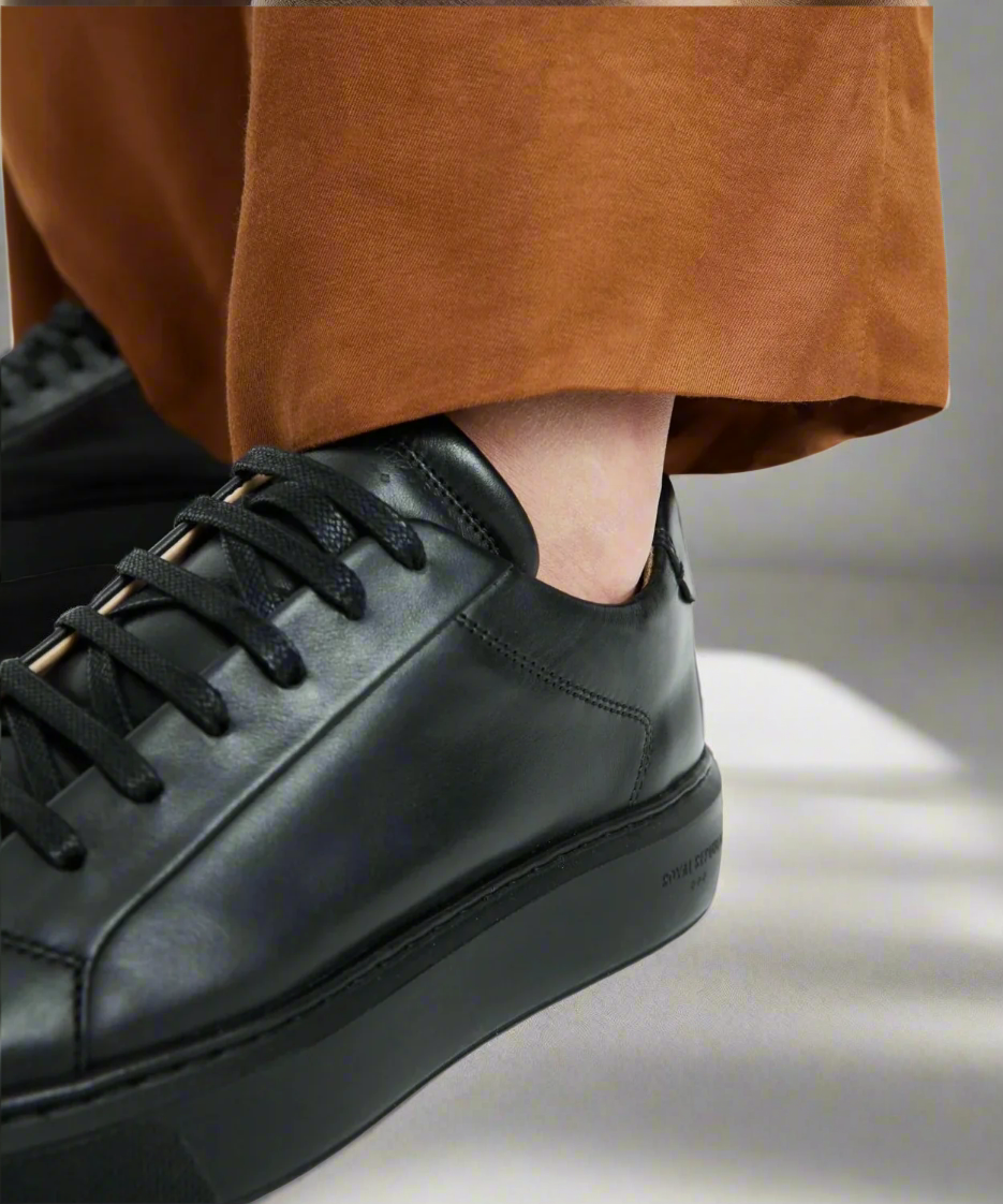 Black Royal RepubliQ leather sneaker worn with a brown garment on a neutral background
