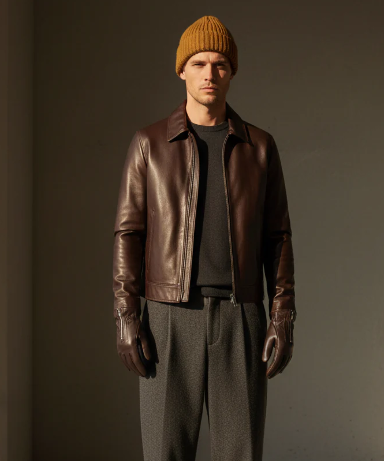 Man wearing a brown leather jacket, mustard beanie, and brown Royal RepubliQ gloves against a dark background