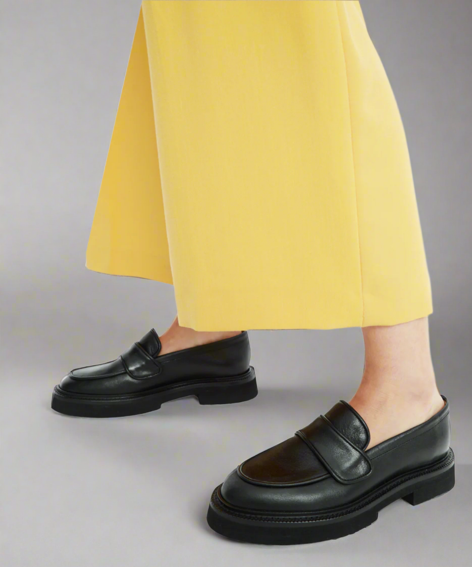 Black loafers worn with yellow pants on a gray background
