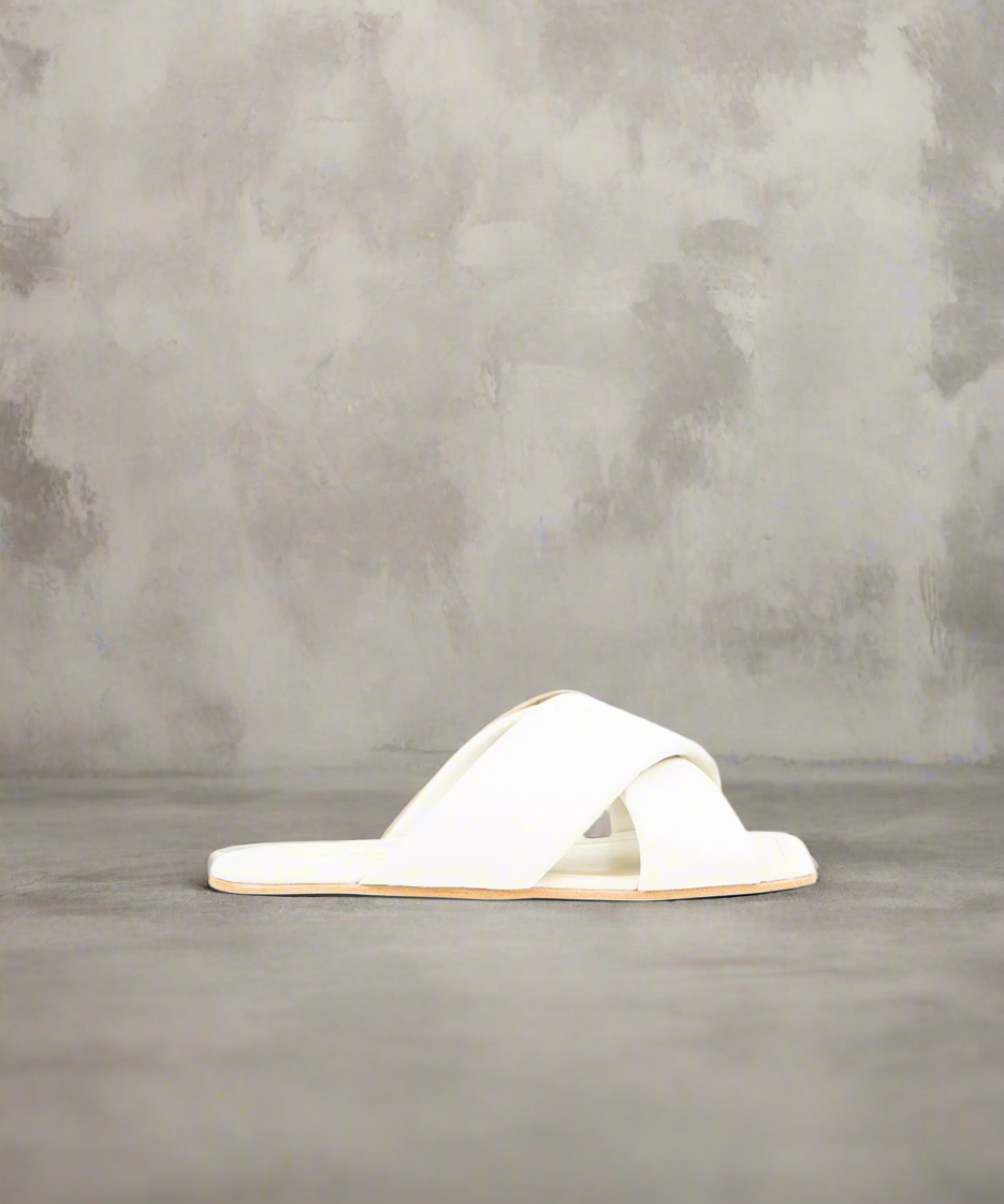 An off white, flat-heeled sandal with a smooth curved toe line, featuring a padded leather strap and cow lining for comfort.