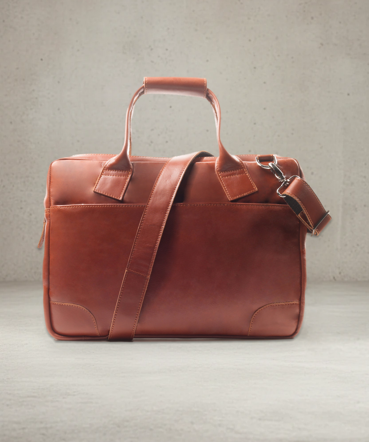 Cognac leather briefcase with top handles and detachable shoulder strap, front view on a concrete studio background