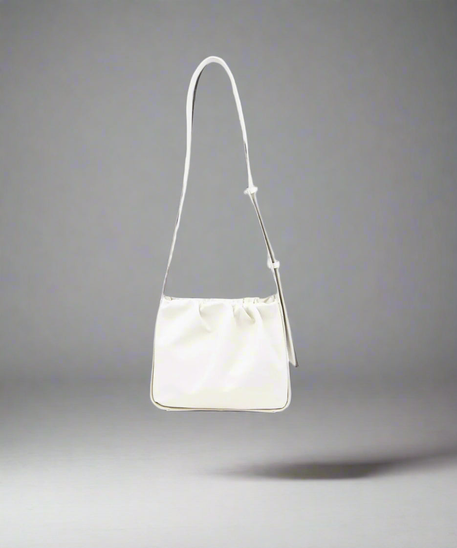An off white crossbody bag made of cow leather with a soft rectangular body and a protective stiff structure around it, featuring gold trimmings and a shoulder strap.