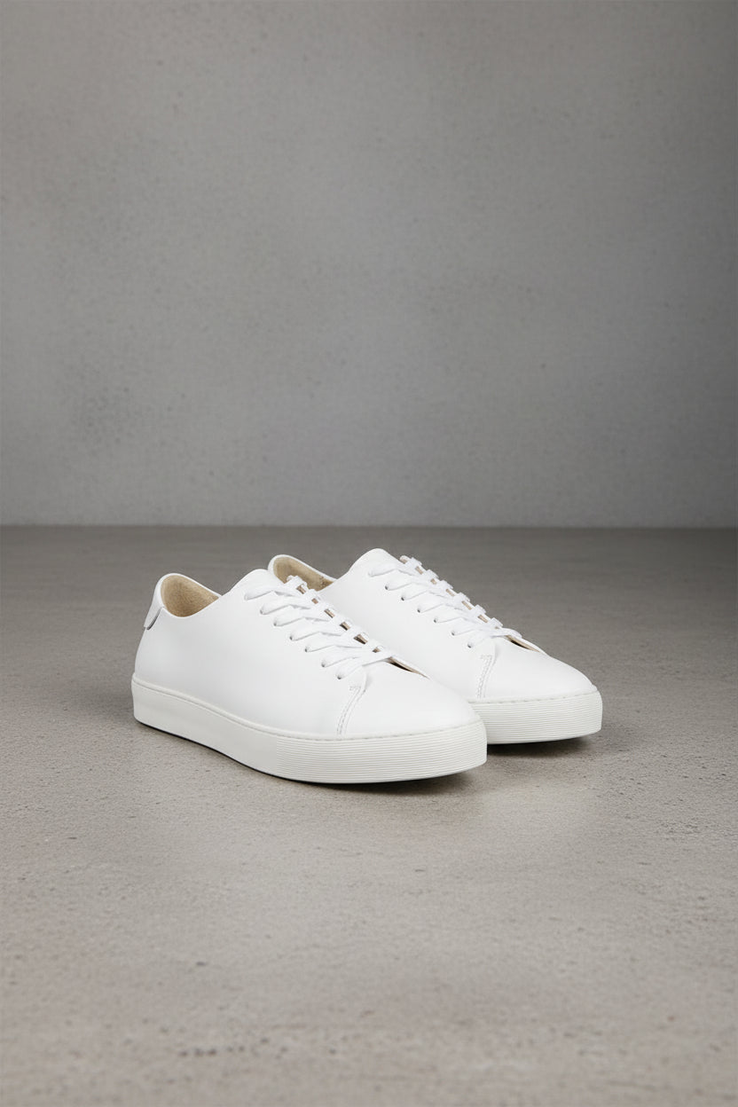 A pair of white leather low-top sneakers with white laces and white rubber soles, displayed on a gray concrete floor against a gray wall.