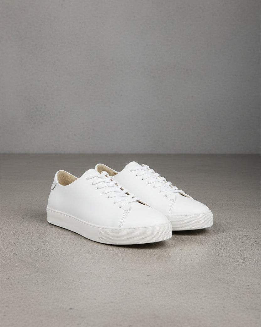 A pair of white leather low-top sneakers with white laces and white rubber soles, displayed on a gray concrete floor against a gray wall.