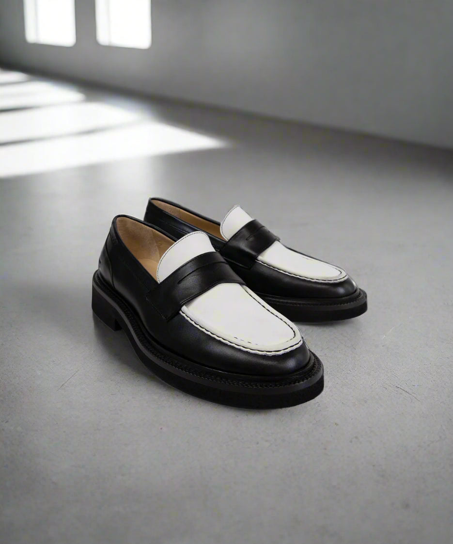 Black and white Royal RepubliQ loafers on a gray floor with sunlight casting shadows.