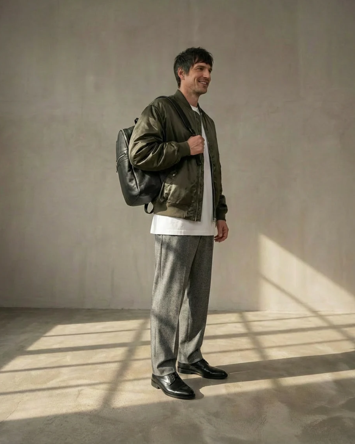 A man wearing a green jacket and gray pants, wearing a black Royal RepubliQ backpack, standing in a room with sunlight casting shadows on the floor.