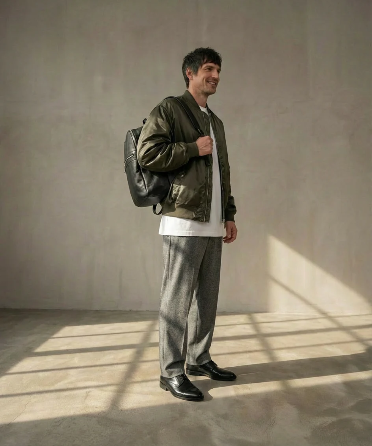 A man wearing a green jacket and gray pants, wearing a black Royal RepubliQ backpack, standing in a room with sunlight casting shadows on the floor.