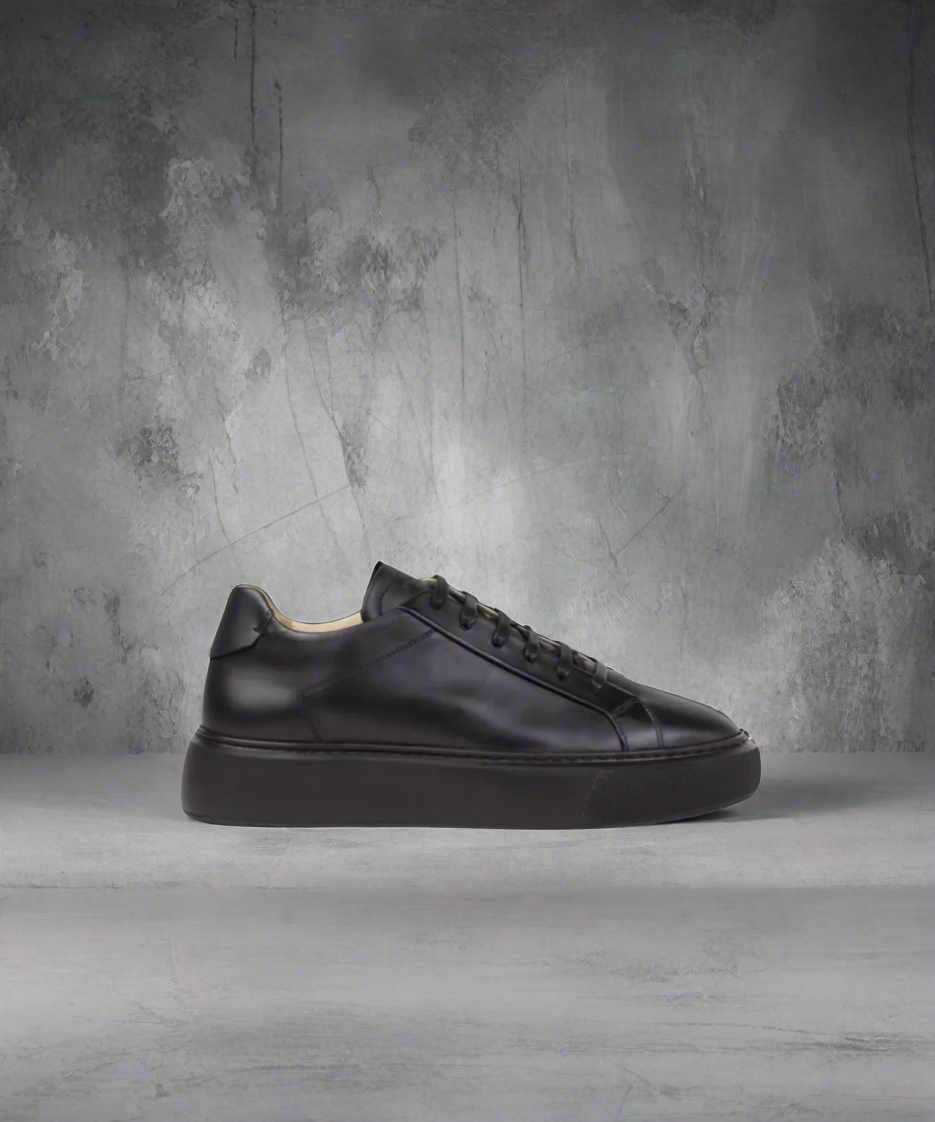 Black Royal RepubliQ leather sneakers on a gray concrete floor with a textured wall background