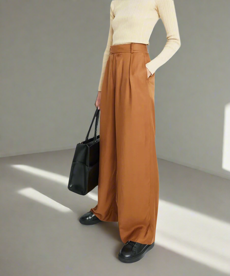 Person wearing brown wide-leg pants and a beige top, holding a black Royal RepubliQ handbag, and black sneakers in a minimalistic setting.