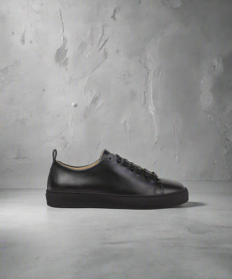 A black low-top men's sneaker with lace-up closure, featuring a smooth leather upper and a flat rubber outsole.