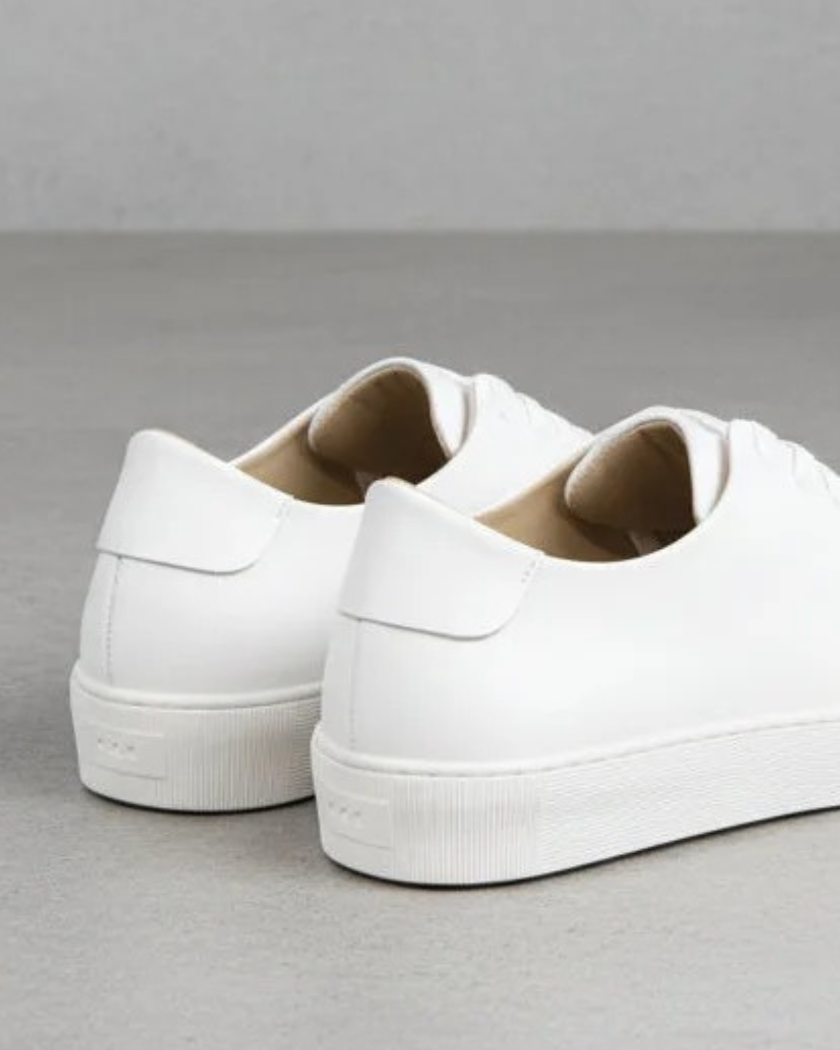 A pair of white leather low-top sneakers shown from the rear three-quarter angle on a neutral gray studio background.