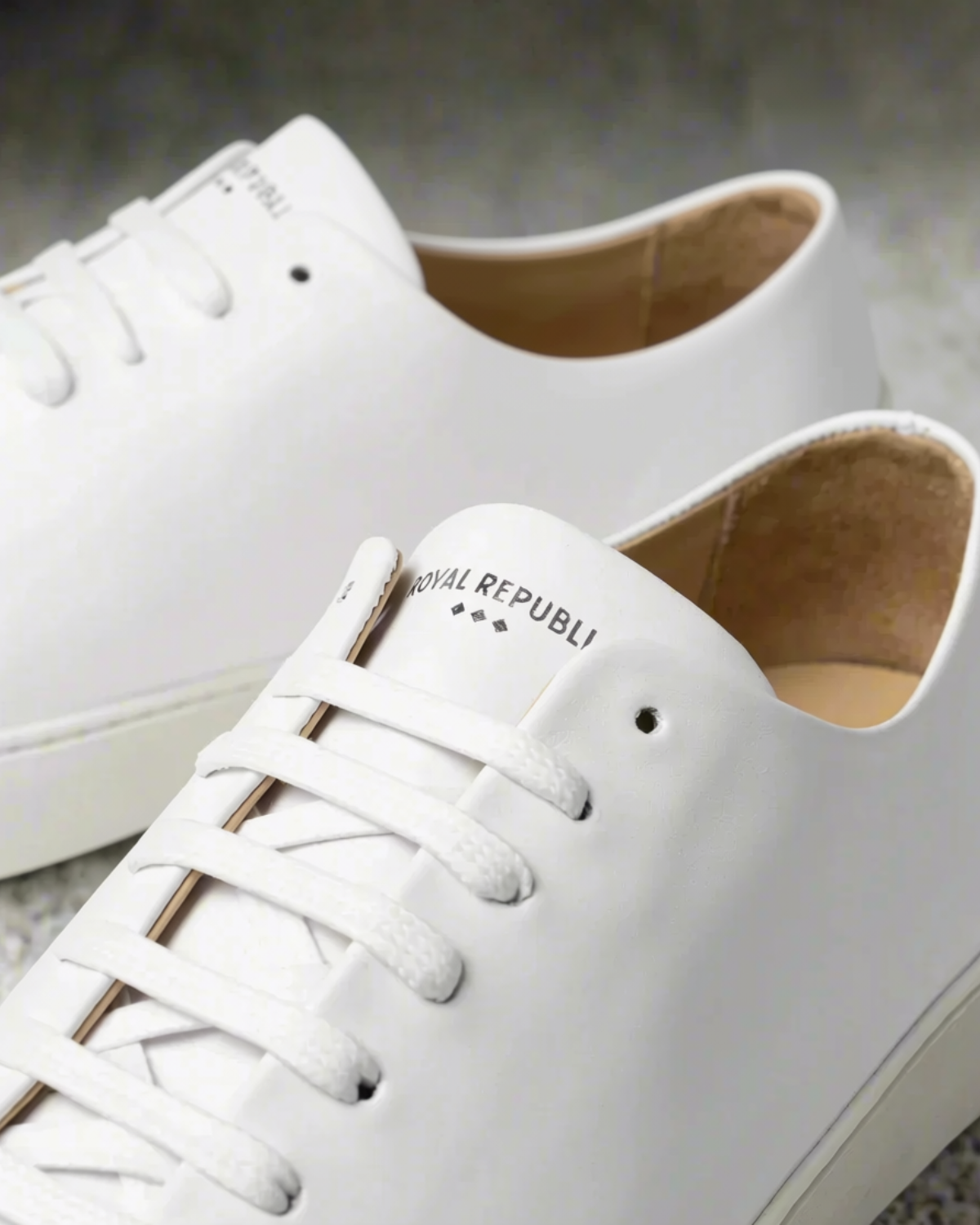 White leather low-top sneakers with white laces and a tan interior, featuring the brand name 'ROYAL REPUBLI' printed on the tongue, shown on a textured gray surface.