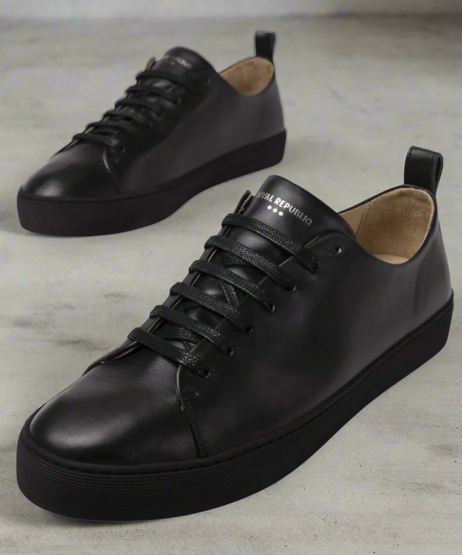 A black Royal RepubliQ leather low-top men's sneaker with lace-up closure, featuring a smooth leather upper and a flat rubber outsole, with brand name on a grey background