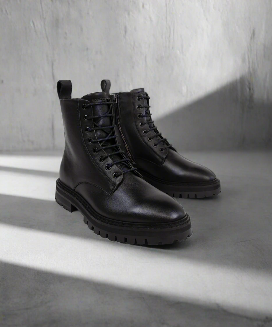 Pair of black leather boots on a gray concrete floor with a light gray wall background.