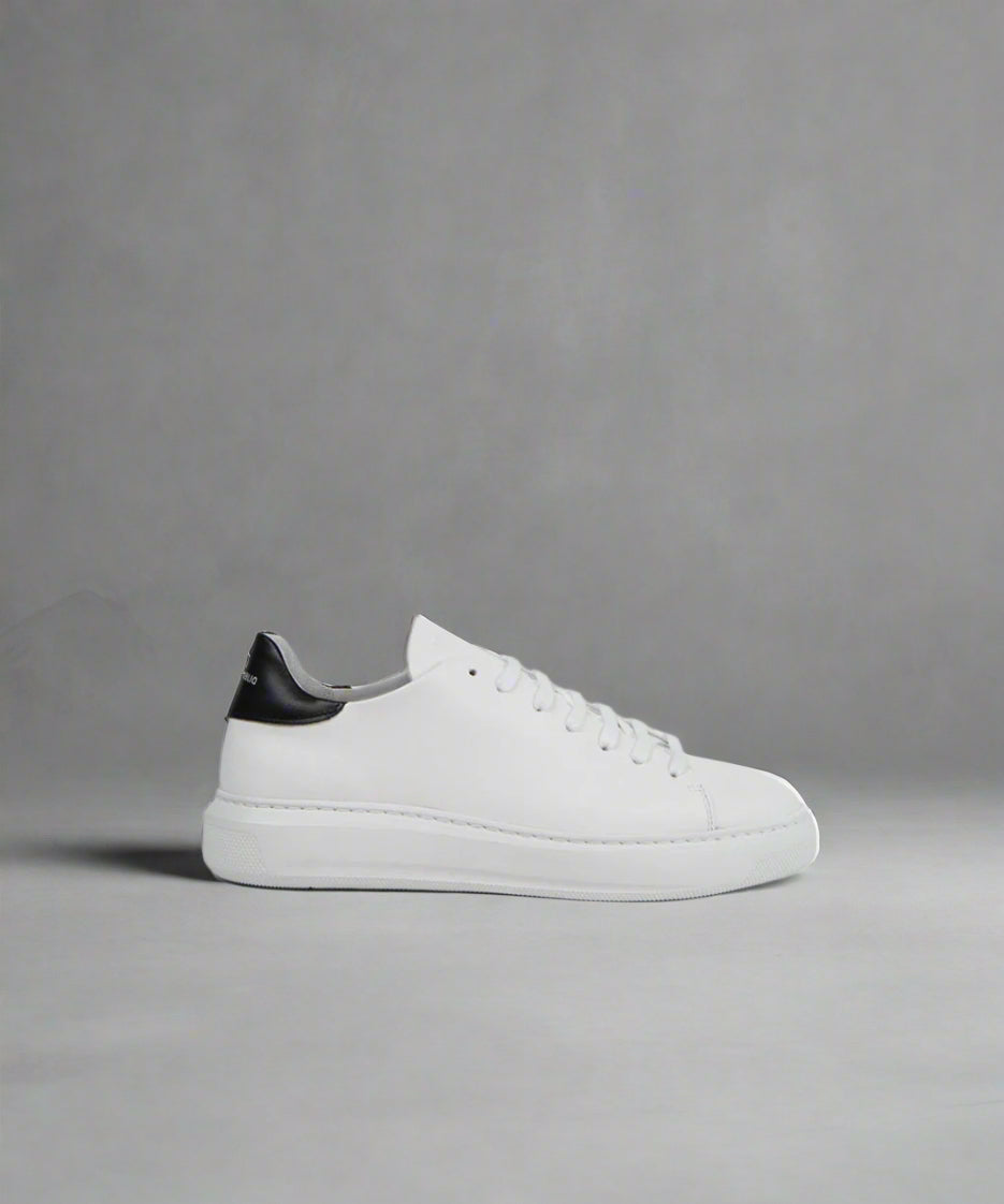 A white low-top sneaker with lace-up closure, featuring a minimalistic design with subtle quilting at the heel and a black detail on the ankle collar.